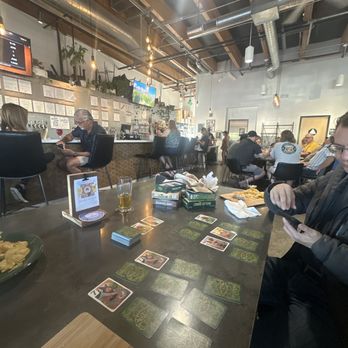 Mindscape Fermentations - The image shows a cafe or bar interior with multiple patrons seated at tables and the bar. Some customers are engaged in a card game on a table, with cards and game boxes visible. The space features an industrial ceiling with exposed ductwork, hanging lights, and artwork on the walls. The bar area has a menu and potted plants, and patrons are using their phones or chatting. The environment appears lively and social. - Breweries Near Me - Rocklin, California