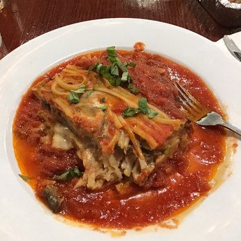 Vinny's Ristorante - A slice of eggplant parmesan with melted cheese, tomato sauce, and chopped basil on top is served on a white plate. The dish is surrounded by additional tomato sauce. The plate is placed on a dark wooden table, with a fork resting on the side and a container of salad in the background. - Italian Near Me - Somerville, Massachusetts