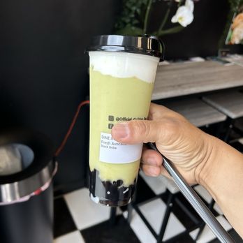 Golden Tea Cafe - Skip lunchFresh Avocado Slush with cheese foam - A hand is holding a tall, clear plastic cup of bubble tea with a black lid. The drink appears to be avocado-flavored with a light green color and has black boba at the bottom. The cup has a label with the flavor details. The background shows a black wall, some floral decorations, and a checkered black-and-white floor, indicating an indoor cafe setting. - Bubble Tea Near Me - Peabody, Massachusetts