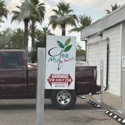 The sign - The image depicts a parking lot with a sign for My Tea & Sweets, a business offering beverages and desserts. The sign includes the business name, logo with leaves, and a notice about tow-away zones. Palm trees are visible in the background, indicating a warm climate. A maroon pickup truck is parked next to the sign, and part of a building with a white tiled exterior is also visible.