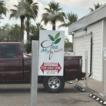 My Tea - The sign - The image depicts a parking lot with a sign for My Tea & Sweets, a business offering beverages and desserts. The sign includes the business name, logo with leaves, and a notice about tow-away zones. Palm trees are visible in the background, indicating a warm climate. A maroon pickup truck is parked next to the sign, and part of a building with a white tiled exterior is also visible. - Bubble Tea Near Me - Phoenix, Arizona
