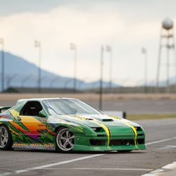 The Tint & Sign Zone - A green Mazda RX-7 with colorful graphics is parked on a racetrack. The car features a large rear wing and aftermarket wheels...