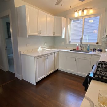 Pearlstone Cutters - The image shows a kitchen with white cabinets and a marble countertop. A window above the sink allows natural light to enter, and a ceiling fan with a light fixture is mounted on the ceiling. The dark hardwood flooring contrasts with the white cabinetry. The kitchen appears clean and modern, with various small items on the countertop near the sink. - Kitchen & Bath Near Me - Chicago, Illinois