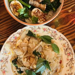 Pad Kee Mao - Two Asian dishes are shown: one with rice, pork belly, boiled egg, and greens, and another with wide rice noodles, vegetables, and basil leaves.