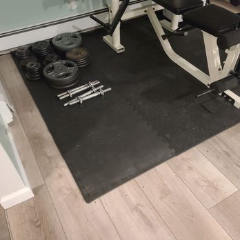 KD Flooring & Tile - Workout room installation with workout mat overtop. - The image shows a corner of a home gym with a black rubber flooring mat. On the mat, there are several weight plates of different sizes, a few dumbbells, and a workout bench with a white frame and black padding. The room has light-colored wooden flooring surrounding the mat, and a wall with a baseboard is visible in the background. - Carpet Installation Near Me