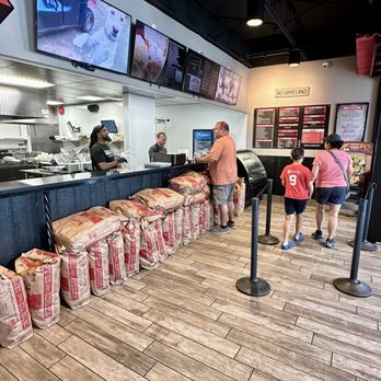 Chaps Pit Beef - The image shows the interior of a Baltimore-style BBQ restaurant with a counter where customers are ordering. Menu screens are mounted above the counter, and staff are preparing food behind it. Several customers, including a man and a woman with two children, are standing in line. The floor is wood-look tile, and bags of supplies are lined up along the front of the counter. The setting appears casual and modern. - Barbeque Near Me - Baltimore, Maryland