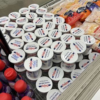 Vons - The image shows numerous small, round containers of Daisy Sour Cream stacked in a refrigerated section of a grocery store. The containers have white lids with the Daisy logo and product information. To the right, there are bags of shredded cheese, and in the lower left corner, bottles of purple-capped condiments are visible. The display is part of a typical grocery store setup, with a metal shelf and a glass front. - Grocery Near Me - Murrieta, California