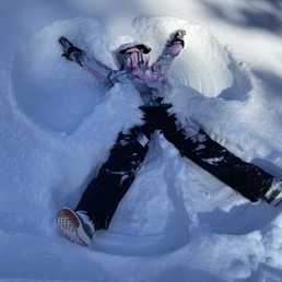 Snow angels are a must! - A child wearing a pink and purple winter snowsuit and black gloves is lying on their back in fresh, white snow, making a snow angel. The child’s arms and legs are spread out, and the snow around them is shaped into the outline of an angel. The background shows snow-covered ground with shadows cast by nearby trees, indicating a sunny day at a ski resort.