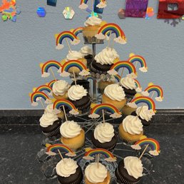 Birthday Cupcake Tower - A tiered cupcake stand holds cupcakes decorated with rainbow toppers, set against a birthday party backdrop with colorful banners and decorations.