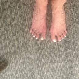 The image depicts a pair of feet with white toenail polish, positioned on a textured, striped surface that appears to be a rug or mat. The feet are close together, and the toes are visible with neatly painted nails. The surface has a pattern of fine, parallel lines in neutral tones. The background includes a small part of a shoe or furniture in the lower left corner.
