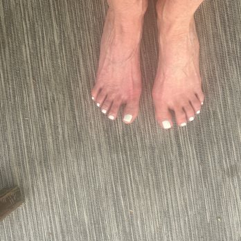 Luxury Nails - The image depicts a pair of feet with white toenail polish, positioned on a textured, striped surface that appears to be a rug or mat. The feet are close together, and the toes are visible with neatly painted nails. The surface has a pattern of fine, parallel lines in neutral tones. The background includes a small part of a shoe or furniture in the lower left corner. - Nail Salons Near Me - Quincy, Massachusetts