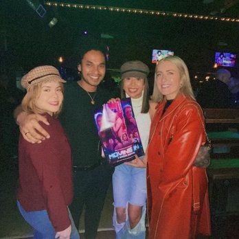 Charlie's Chicago - A group of four women and one man stand together in a dark, indoor setting, holding a poster with vibrant colors, possibly at a nightclub or event. - Gay Bars Near Me - Chicago, Illinois