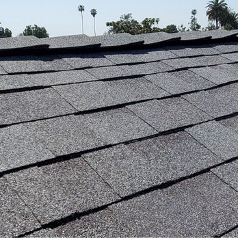 GreenCal Construction - Roofing 2021 - The image depicts a roof with asphalt shingles arranged in overlapping rows, featuring a vent on the right side. The background includes trees, palm trees, and a clear sky, indicating an outdoor setting. - Roofing Near Me - Bellflower, California