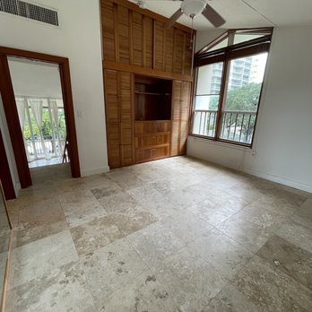 Pure Moving - Bedroom - An empty room with a tiled beige floor, large windows with wooden frames providing natural light and a view of nearby buildings, and a wooden closet with louvered doors and built-in shelving. A ceiling fan is installed, and a doorway leads to another space with a balcony or outdoor area visible through the adjacent room. - Movers Near Me - Miami, Florida