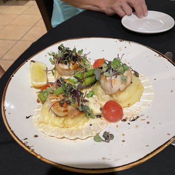 The Front Yard - JUMBO SCALLOPS IN THE SHELL - The image shows a seafood dish with three scallops, each topped with microgreens, served on a large, round, decorative plate. The dish includes vegetables such as cherry tomatoes and asparagus, with lemon wedges on the side. The presentation is arranged on a black table in a restaurant setting, with a person in a light blue shirt visible in the background. - Mediterranean Near Me - Ogunquit, Maine