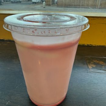 Snarfburger - Strawberry malted milkshake - A pink strawberry malted milkshake is served in a clear plastic cup with a sealed lid. The cup is placed on a dark wooden surface near a window, with a view of a residential neighborhood outside. The background shows houses, trees, and parked cars, indicating an indoor setting with natural light. - Burgers Near Me - Boulder, Colorado
