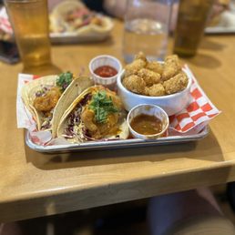 Fish Tacos - A metal tray holds fish tacos with mango salsa, accompanied by tater tots and small cups of salsa and dipping sauce, on a wooden table.