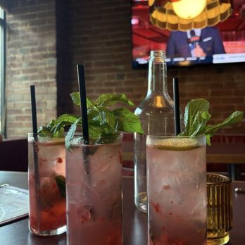 Cherry's High Dive - Raspberry Mojitos - Three raspberry mojitos with fresh mint leaves and ice are placed on a table in a restaurant. A glass bottle is positioned behind the drinks, and a yellow candle holder is visible to the right. In the background, a television screen displays a news broadcast, and a stained glass-style hanging lamp is above the drinks. The restaurant has brick walls and a window on the left side. - Dive Bars Near Me - Toronto, Ontario
