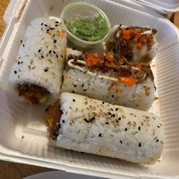 A takeout container holds three sushi rolls topped with sesame seeds, with a small cup of green sauce in the center. The sushi appears to contain various fillings, and the container is placed on a wooden table. Additional takeout boxes are visible in the background.