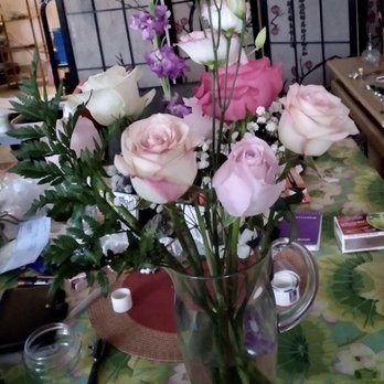 Happy Flowers - Mother's Day flowers charged $65, should be half that price - A bouquet of mixed flowers, including pink and white roses, purple flowers, and green foliage, arranged in a clear glass pitcher. The table has a colorful, floral-patterned tablecloth and various objects around, such as jars, papers, and small boxes. The background includes a decorative black metal screen and other household items. - Florists Near Me - Flushing, New York