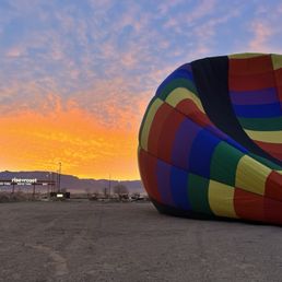 A multicolored hot air balloon is deflated on the ground near a desert area during sunset, with a restaurant and mountains in the background.