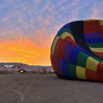 Fly Albuquerque - A multicolored hot air balloon is deflated on the ground near a desert area during sunset, with a restaurant and mountains in the background. - Hot Air Balloons Near Me - Albuquerque, New Mexico