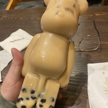 Royaltea - A bear-shaped plastic bottle filled with bubble tea, featuring black tapioca pearls at the bottom, is held over a wooden table in a cafe setting. - Bubble Tea Near Me - Toronto, Ontario