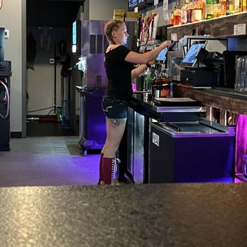 The Vixen - a woman at a bar - A woman with braided hair, tattoos on her thigh, and red lace-up boots is standing behind a bar counter, operating a touchscreen device. The bar area features liquor bottles on shelves, multiple screens mounted on the wall, and purple lighting accents. The setting appears to be a nightclub or bar with a modern, illuminated ambiance. - Music Venues Near Me - McHenry, Illinois