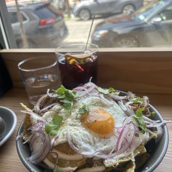 Downstate - A bowl containing a Mexican-style dish with fried eggs, sliced red onions, and cilantro on top, placed on a wooden table near a window. Behind the bowl, there is a glass of iced beverage with a straw and a slice of lemon, along with a glass of water. The window shows parked cars and a street scene outside, with buildings and vehicles visible in the background. - Cafes Near Me - Newburgh, New York