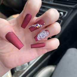 A hand with long, matte burgundy nails features rhinestone embellishments and a 3D white flower nail art design, set against a car interior background.