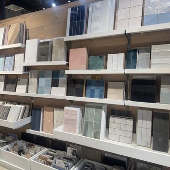 Bodega Style - The image shows a retail showroom with shelves filled with different types of tiles and surface materials. The tiles are arranged in organized sections, featuring various colors, patterns, and textures suitable for home improvement projects. The display includes large format tiles, smaller mosaic pieces, and sample boards, with lighting overhead highlighting the products. The setting appears to be a dedicated space for interior design and renovation materials. - Building Supplies Near Me - Anaheim, California