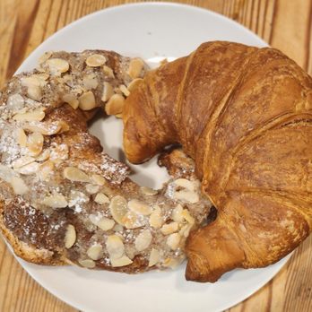 Cafe Barista - Suffield - pecan horn and regular croissant - A plain croissant and an almond croissant with sliced almonds on top are placed on a white plate, set on a wooden surface. - Coffee & Tea Near Me - Suffield, Connecticut