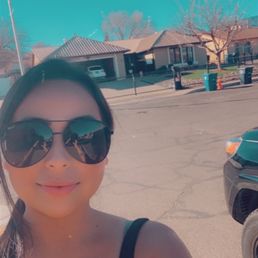 A woman with dark hair and sunglasses is taking a selfie outside in a residential area with houses and parked cars.