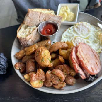 VUE Rooftop - A breakfast plate featuring toasted bread, sausage links, roasted potatoes, fried eggs, and a small dish of butter, served on a dark table. - New American Near Me - Washington, DC, District of Columbia
