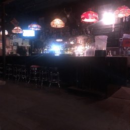 The image shows a dimly lit bar with a dark counter and several metal stools lined up in front. Hanging vintage-style lamps provide ambient lighting, and the back bar is stocked with bottles and glassware. The overall atmosphere appears cozy and retro, with various signs and decorations on the walls.