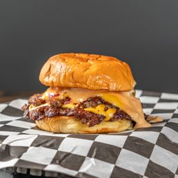 Son of a Bun - Double - A cheeseburger with a beef patty, melted cheese, and a toasted bun is placed on black-and-white checkered paper, with a dark background.