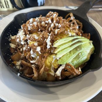 Hash Kitchen Arcadia - Carnitas Hash - A black skillet holds Chile Relleno Hash, featuring diced potatoes, topped with shredded cheese, crispy fried onion strings, and sliced avocado. The dish is served on a white plate, with a coffee mug and other breakfast items visible in the background, indicating a brunch setting. - Breakfast & Brunch Near Me - Phoenix, Arizona