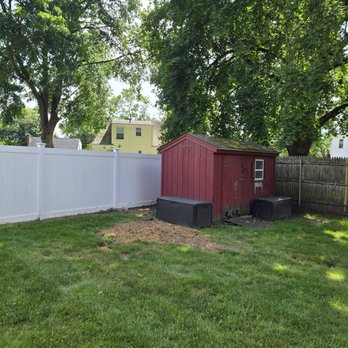 deras tree service - The image shows a backyard with a red wooden shed positioned near a white privacy fence. The yard has green grass with a small patch of dirt in front of the shed. Large trees with dense foliage are visible in the background, providing shade and a natural setting. The scene appears to be taken on a sunny day with clear skies. - Tree Services Near Me - Pawtucket, Rhode Island