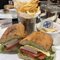 Southwest Turkey - A meal featuring a sandwich with sliced turkey, lettuce, tomato, and cheese on toasted baguette bread. The sandwich has small flags with logos inserted into each half. It is served with a side of French fries in a metal cup lined with paper and a bowl of coleslaw containing shredded vegetables and corn. The setting appears to be a casual dining restaurant with glasses of water and other dishes visible in the background.