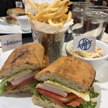 Silver Diner - Southwest Turkey - A meal featuring a sandwich with sliced turkey, lettuce, tomato, and cheese on toasted baguette bread. The sandwich has small flags with logos inserted into each half. It is served with a side of French fries in a metal cup lined with paper and a bowl of coleslaw containing shredded vegetables and corn. The setting appears to be a casual dining restaurant with glasses of water and other dishes visible in the background. - Diners Near Me - McLean, Virginia