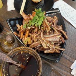 pig ear - Sliced pig ears are presented on a black square plate, garnished with cilantro and sliced green onions, accompanied by soy sauce and a dipping sauce.