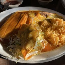 #1 Cheese Enchilada, tamales and taco - A white oval plate contains a tamale covered in sauce, a taco with shredded lettuce and cheese, and a portion of Mexican rice. The dish is served on a dark table with small bowls of sauce and other dishes partially visible around it.