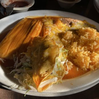 Chalo's - #1 Cheese Enchilada, tamales and taco - A white oval plate contains a tamale covered in sauce, a taco with shredded lettuce and cheese, and a portion of Mexican rice. The dish is served on a dark table with small bowls of sauce and other dishes partially visible around it. - Mexican Near Me - Globe, Arizona