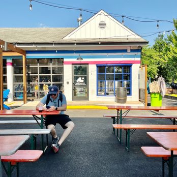 Aslin Coffee - A person wearing a cap and sunglasses sits at an outdoor picnic table, using a phone, in front of a building with colorful stripes and large windows. - Coffee & Tea Near Me - Washington, DC, District of Columbia