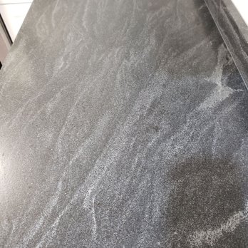 Montes Marble & Granite - A photograph shows a dark gray granite countertop with a textured, speckled surface. The surface has a natural stone appearance with fine white and gray mineral deposits, creating a speckled pattern. The countertop edges are visible, and part of a kitchen setting can be seen in the background, including white cabinetry. The surface appears smooth and polished, suitable for kitchen or bathroom use. - Building Supplies Near Me - Atkinson, New Hampshire