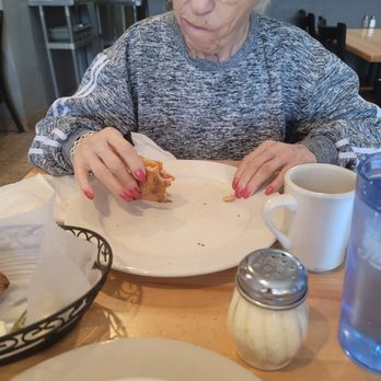 Pizzeria Bacio - An elderly woman with glasses and gray hair, dressed in a gray sweater with white stripes on the sleeves, sits at a wooden table in a restaurant. She is holding a piece of food in her right hand and appears to be looking at her plate. The table has a white mug, a water glass, a small shaker, and a basket of bread or fried food. The background shows shelves with dishes and a dining area with chairs and tables. - Pizza Near Me - Poughkeepsie, New York