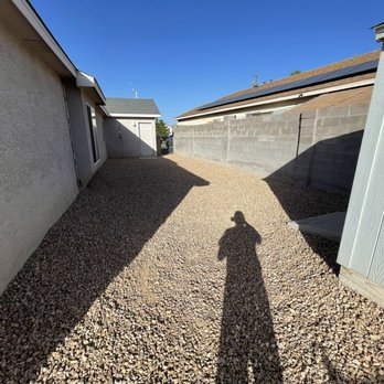 Empire Yard Maintenance - a shadow of a person standing in a driveway - A backyard area covered with small gravel stones, bordered by side walls on both sides. The left wall is part of a house, and the right wall is a fence or wall of another structure. The shadow of the person taking the photo is cast on the gravel, indicating sunlight from the front. The background shows a small shed or outbuilding and neighboring rooftops under a clear blue sky. - Landscaping Near Me
