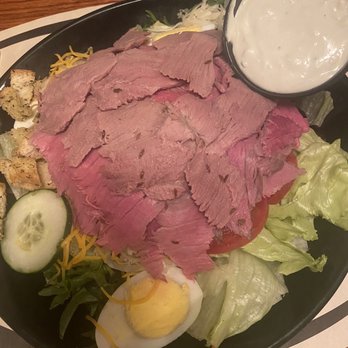 Beef'n Barrel - Beef salad - A beef salad served on a black plate includes sliced roast beef placed over shredded lettuce, with cucumber slices, a halved boiled egg, shredded cheese, and croutons. A small cup of creamy dressing is positioned at the top right of the plate. The dish is presented on a wooden table, with a person wearing a blue shirt visible in the background. - Steakhouses Near Me - Olean, New York