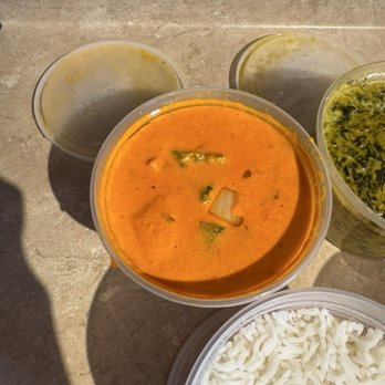 Shalimar - Chicken Tikka Masala - Several plastic containers of Indian food are arranged on a stone surface, including a bowl of orange curry with vegetables, a bowl of cooked rice, and other side dishes with green and yellow sauces. The food appears freshly prepared and is served in takeout containers. - Indian Near Me - Delmar, New York