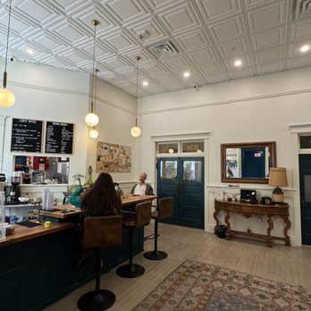 Earl's Cafe - Coffee shop in lobby of hotel - The image shows the interior of a coffee shop with a high, patterned ceiling and pendant lighting. There are three people seated at the bar, with one person facing away and two facing the barista. The counter has coffee-making equipment and a menu board. The wall behind the counter displays a collage and a coffee menu. To the right, there is a dark green door with a sign above it, a mirror, and a wooden side table with decorative items and a lamp. The floor has a patterned rug, and the overall decor combines modern and vintage elements. - Coffee & Tea Near Me - Bisbee, Arizona